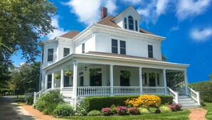 A two-story white house with a porch, surrounded by well-maintained gardens and a clear blue sky. - Fordham House (Greenport)
