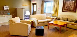 A hotel room with a beige sofa set, a wooden coffee table, a black ottoman, a small table with a lamp, and a framed picture on the wall. - Lodging on the Square (Berlin)