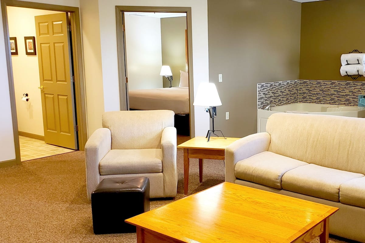 A hotel room with a beige sofa, two armchairs, a wooden coffee table, a small ottoman, a bathroom with a bathtub, and a mirror.