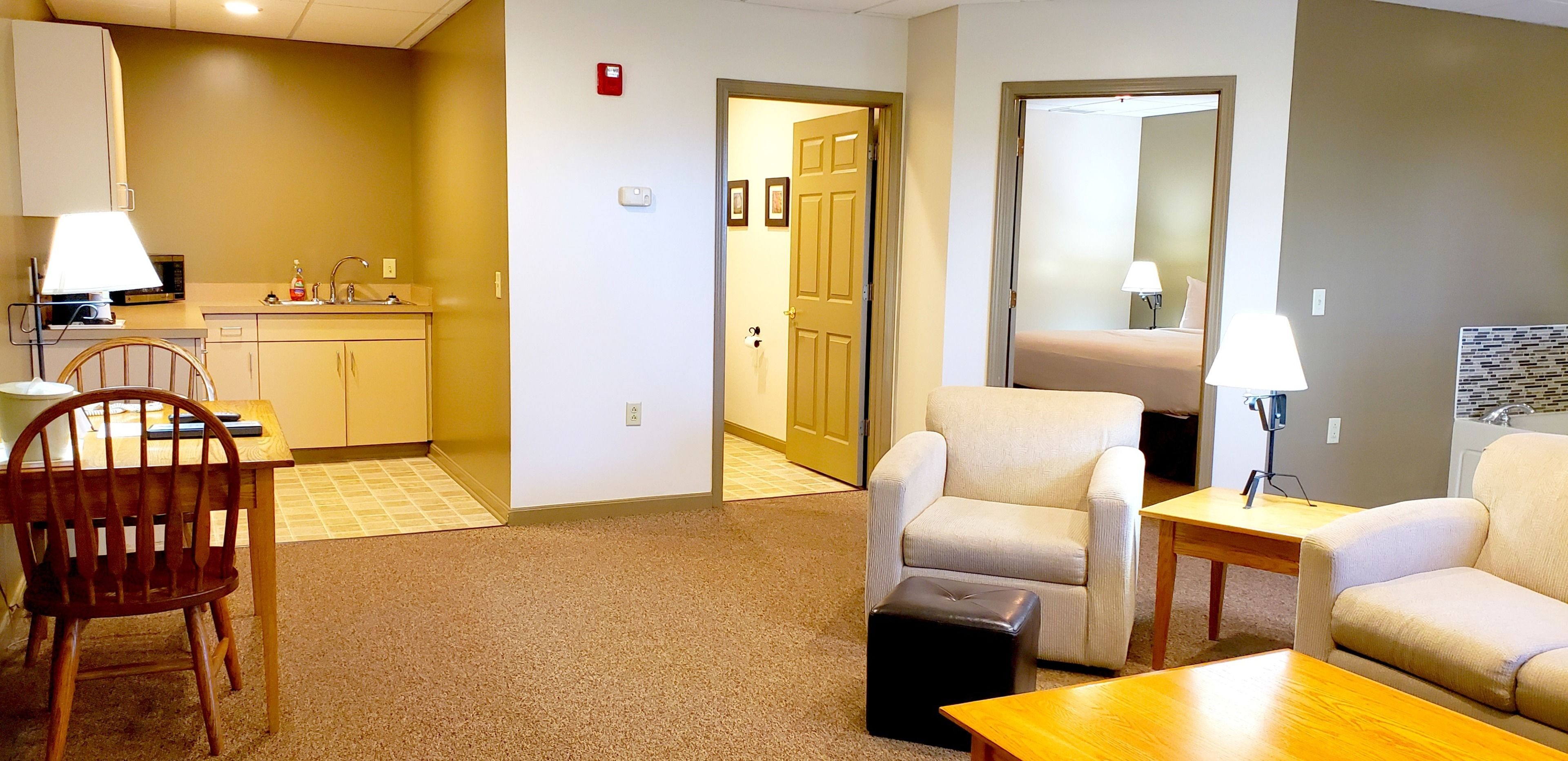 A hotel room with a kitchenette, a dining area with a wooden table and chairs, a sofa, and a small ottoman.