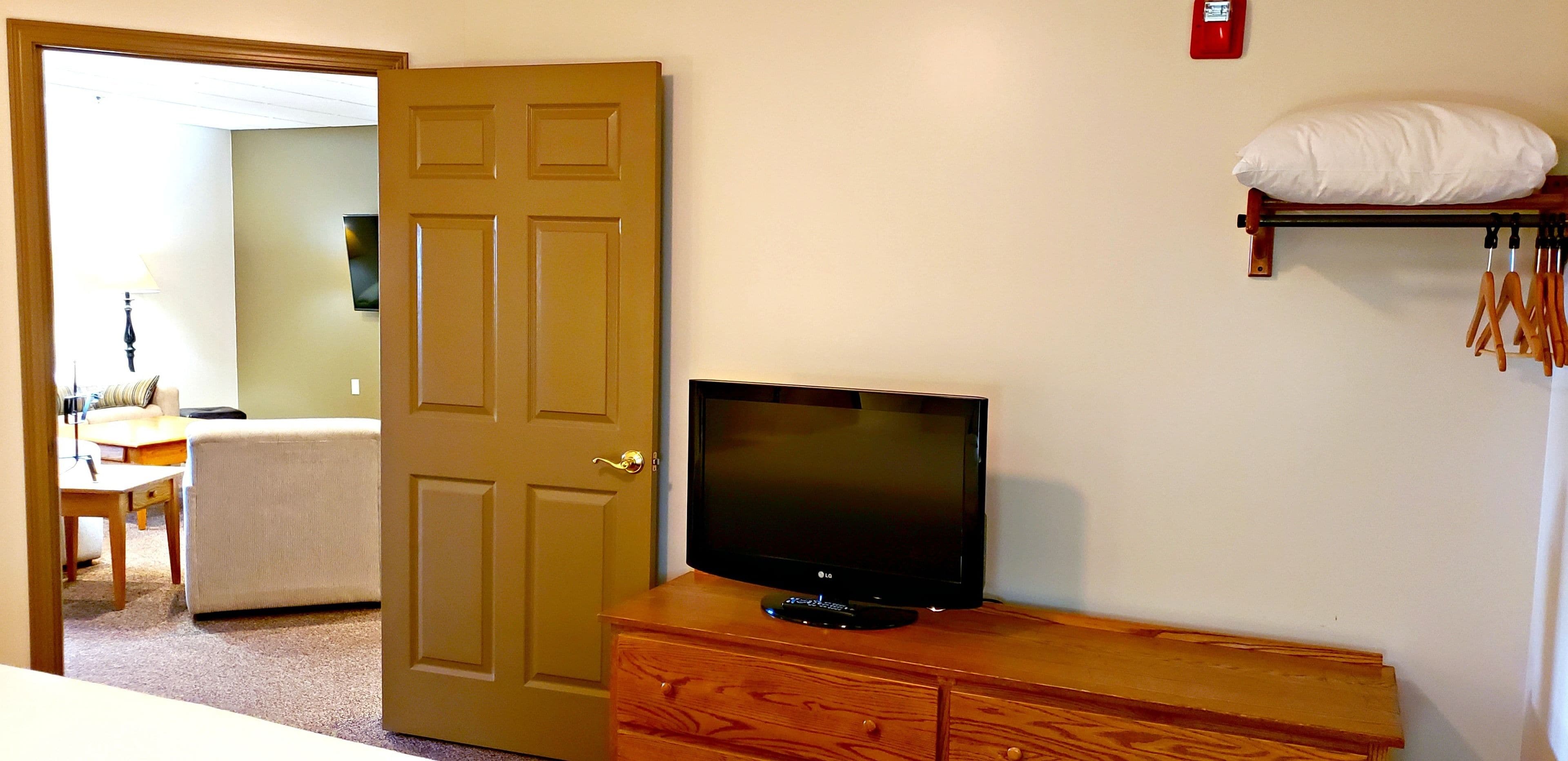 A room with a wooden dresser, a television on top, a door, and a shelf with a pillow and some items.