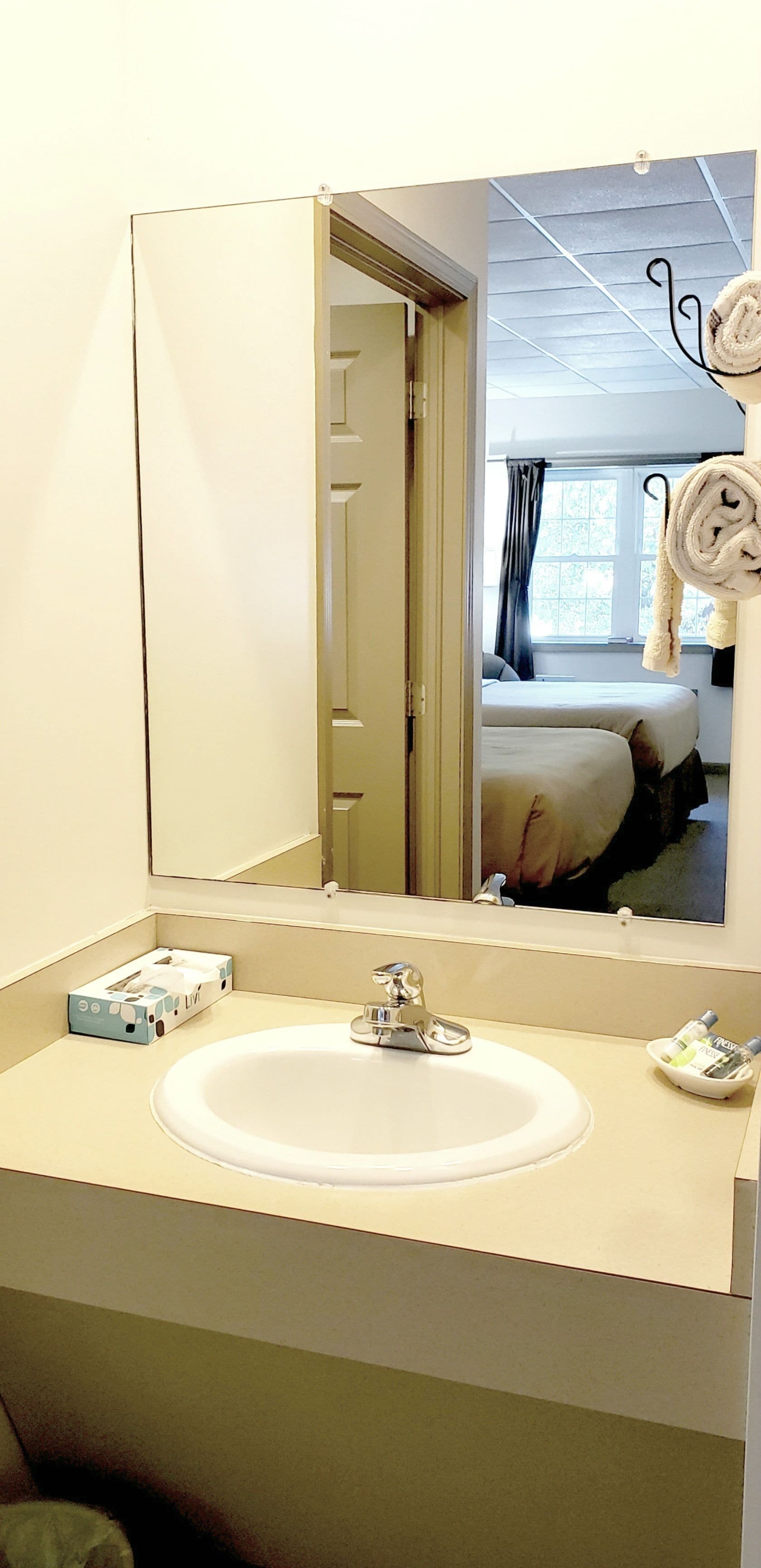 A hotel bathroom with a sink, mirror, and a view of a bedroom with a bed and a window.