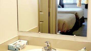 A hotel bathroom with a sink, mirror, and a view of a bedroom with a bed and a window.