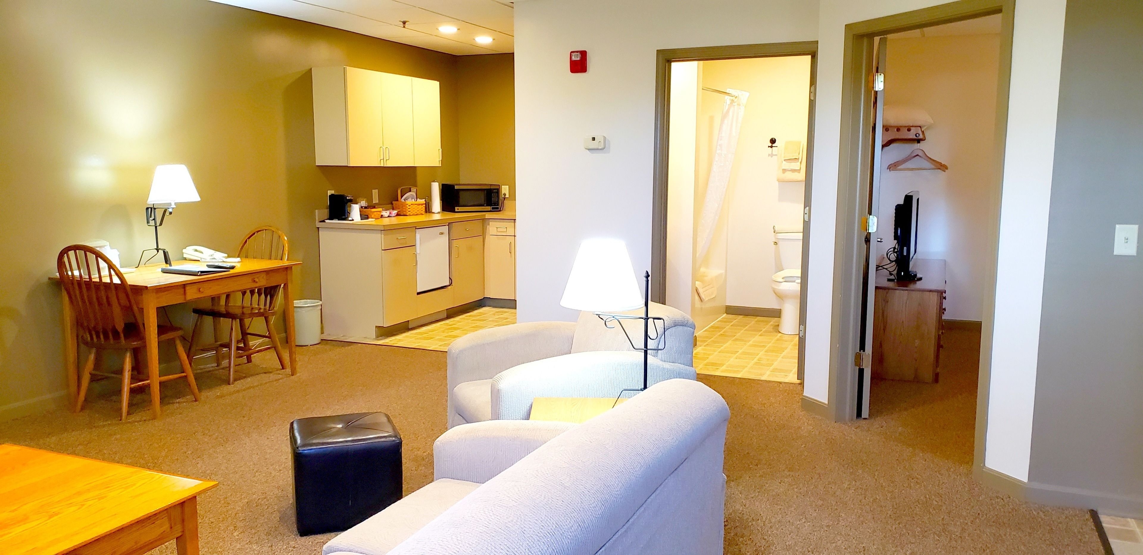 Habitación de hotel compacta con kitchenette, comedor con mesa y sillas, sofá y un pequeño otoman.