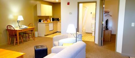 Habitación de hotel compacta con kitchenette, comedor con mesa y sillas, sofá y un pequeño otoman.