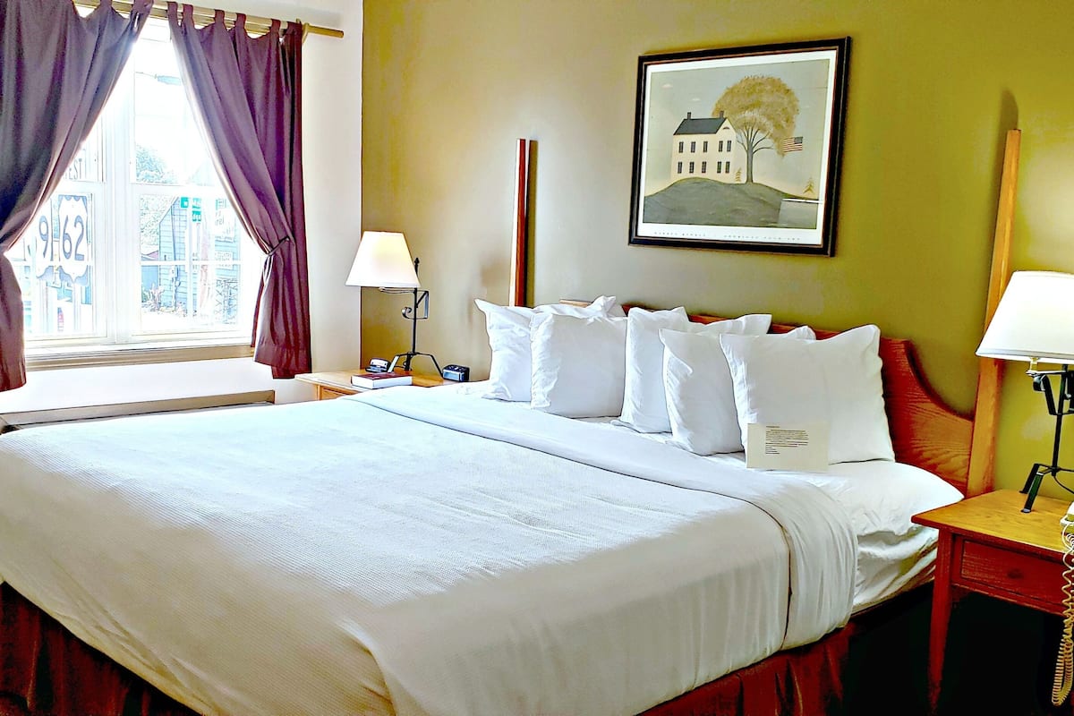 A hotel room with a large bed, two bedside tables, a window with curtains, a lamp, a framed picture on the wall, and a telephone on the nightstand.