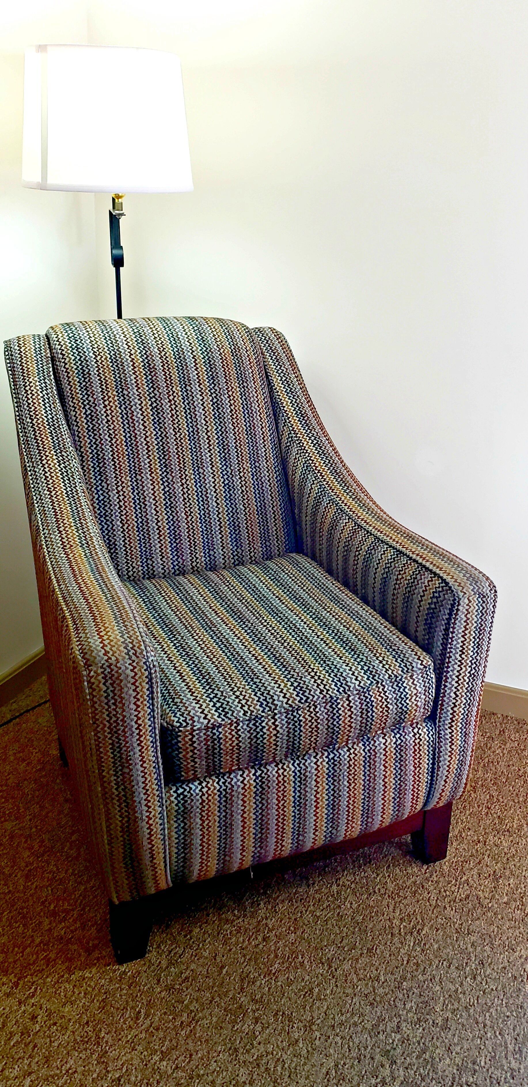 A chair with a woven pattern, positioned in a room with a carpeted floor and a lamp in the background.