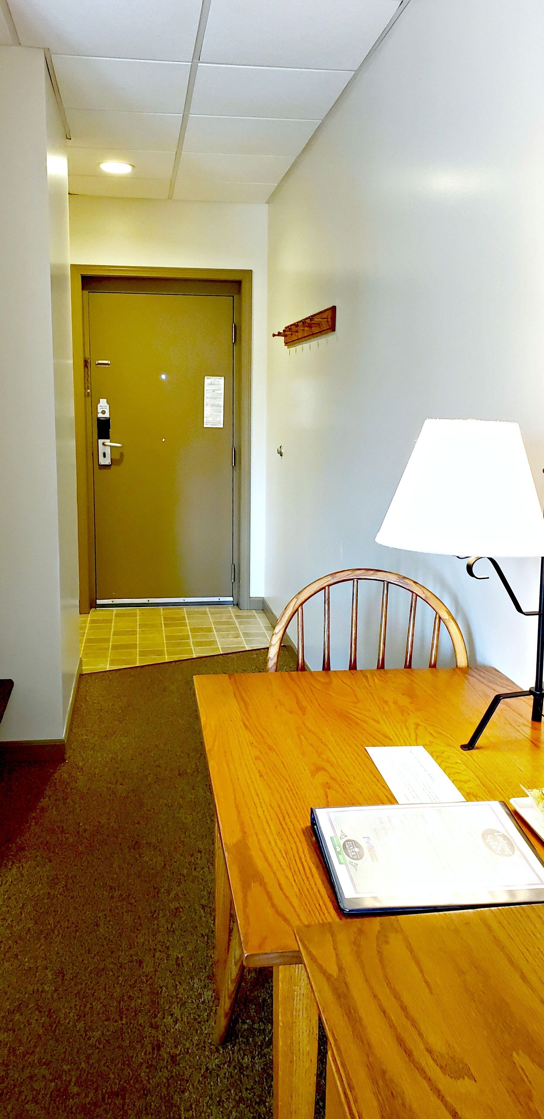 Una habitación de hotel con una mesa de madera, una silla, una lámpara y una puerta que da a otra estancia.