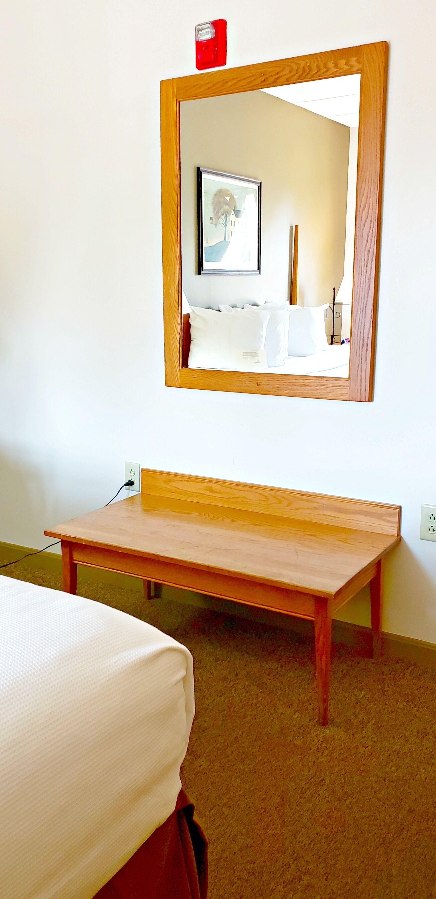 A hotel room with a wooden bench, a bed with white bedding, a large mirror, and a wall socket.