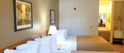 Ein Hotelzimmer mit zwei Betten, einem Nachttisch, einer Lampe, einem gerahmten Bild und einer kleinen Küchenzeile.
