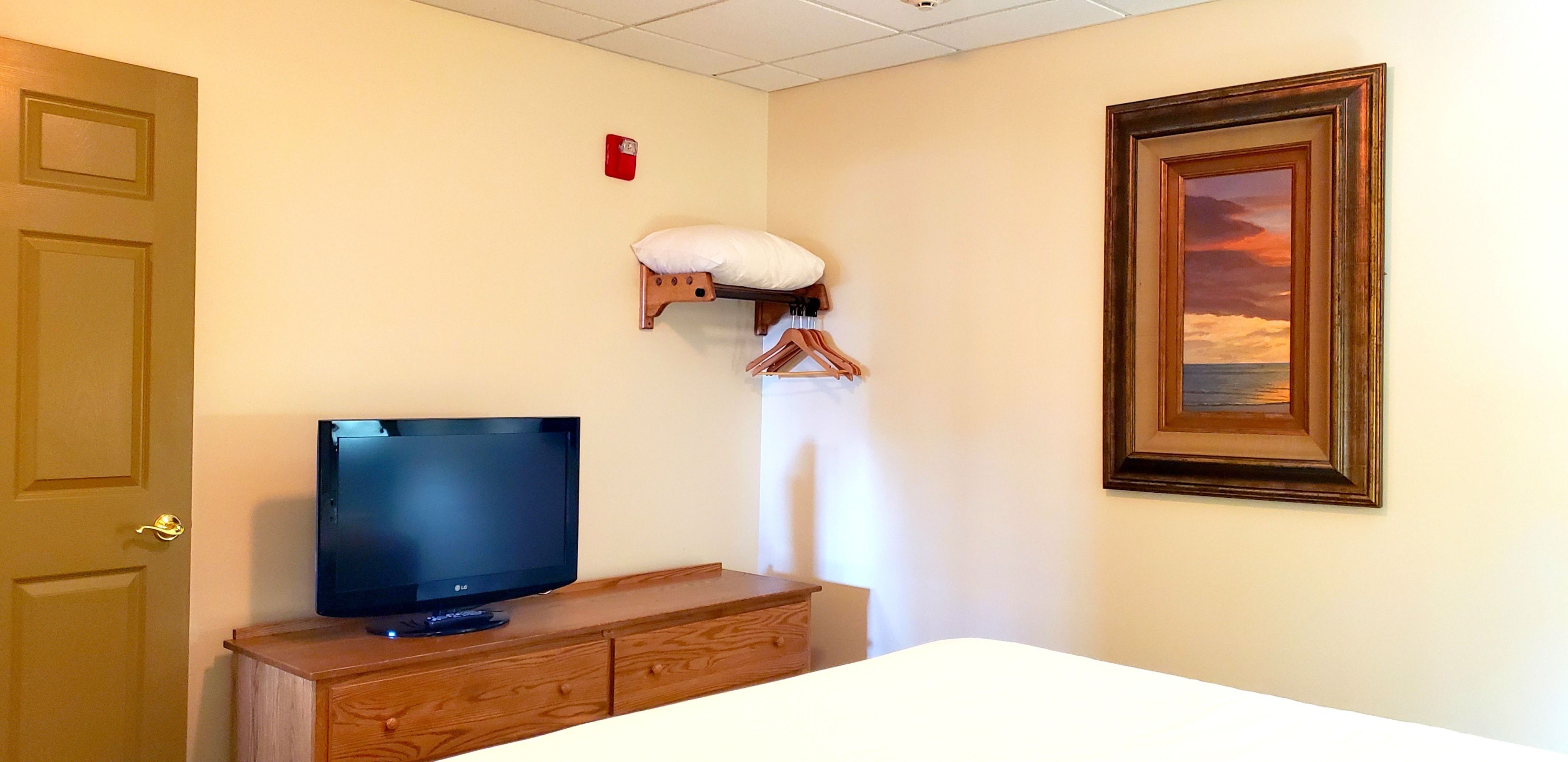 Une chambre d’hôtel avec une télévision posée sur une commode en bois, un oreiller blanc sur une étagère et un tableau encadré accroché au mur.