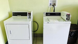Two front-loading washing machines in a laundry room with tiled flooring and a trash can. - Greystone Motel (Hillsboro)