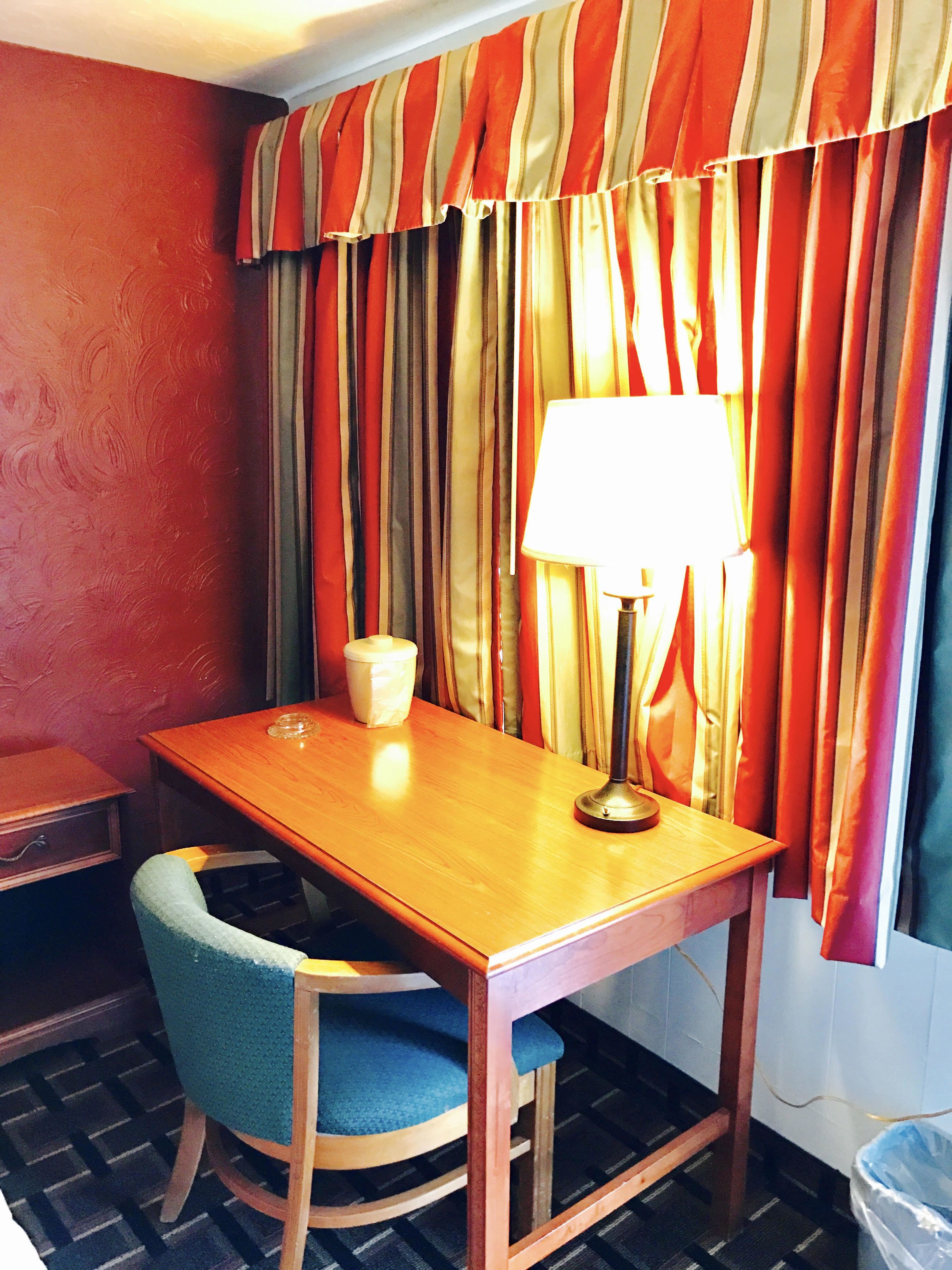 A small, simple room with a wooden table, a blue chair, a lamp, and a window with striped curtains.