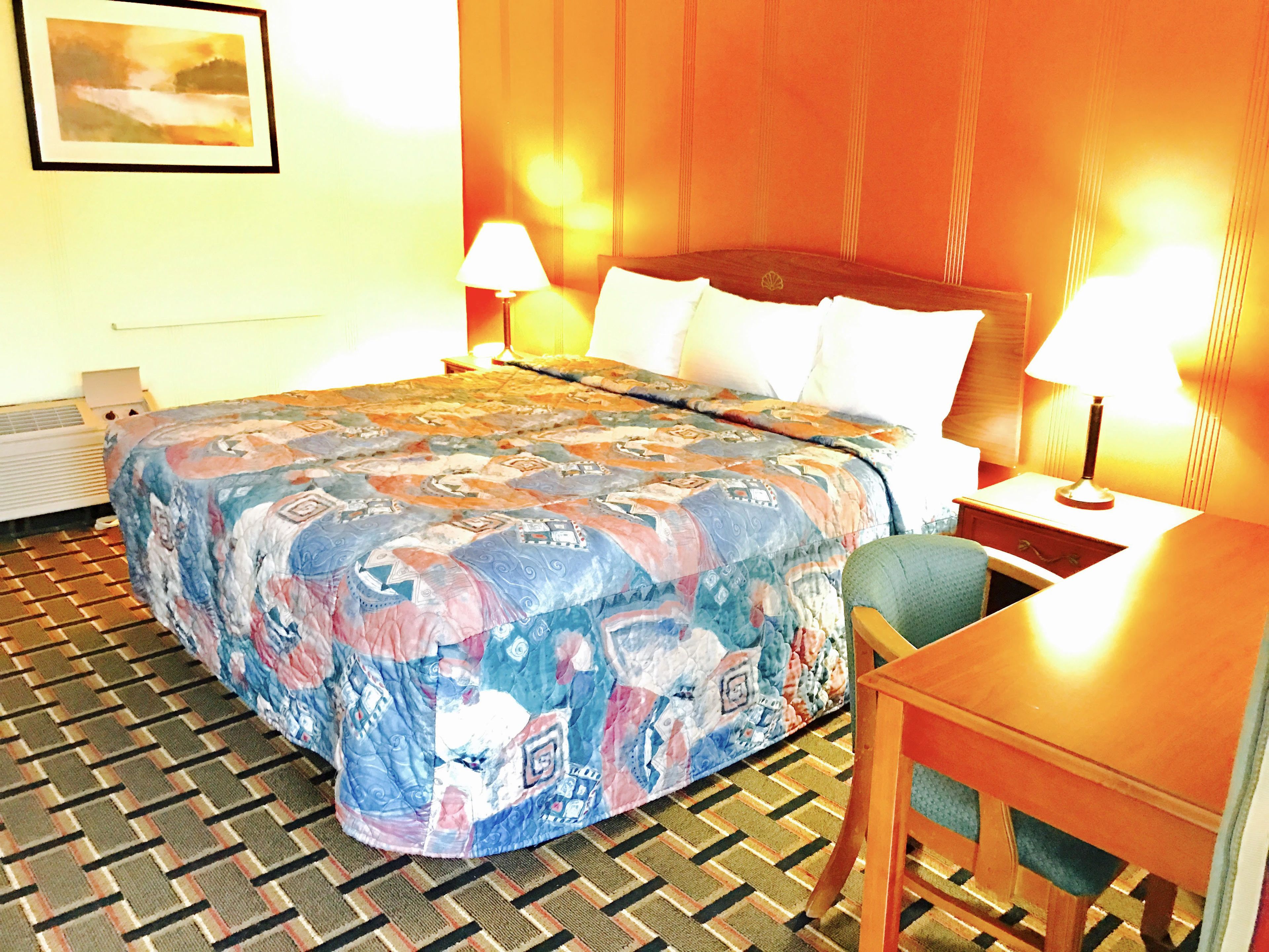 A hotel room with a large bed, two bedside lamps, a wooden headboard, a small table, and a chair.