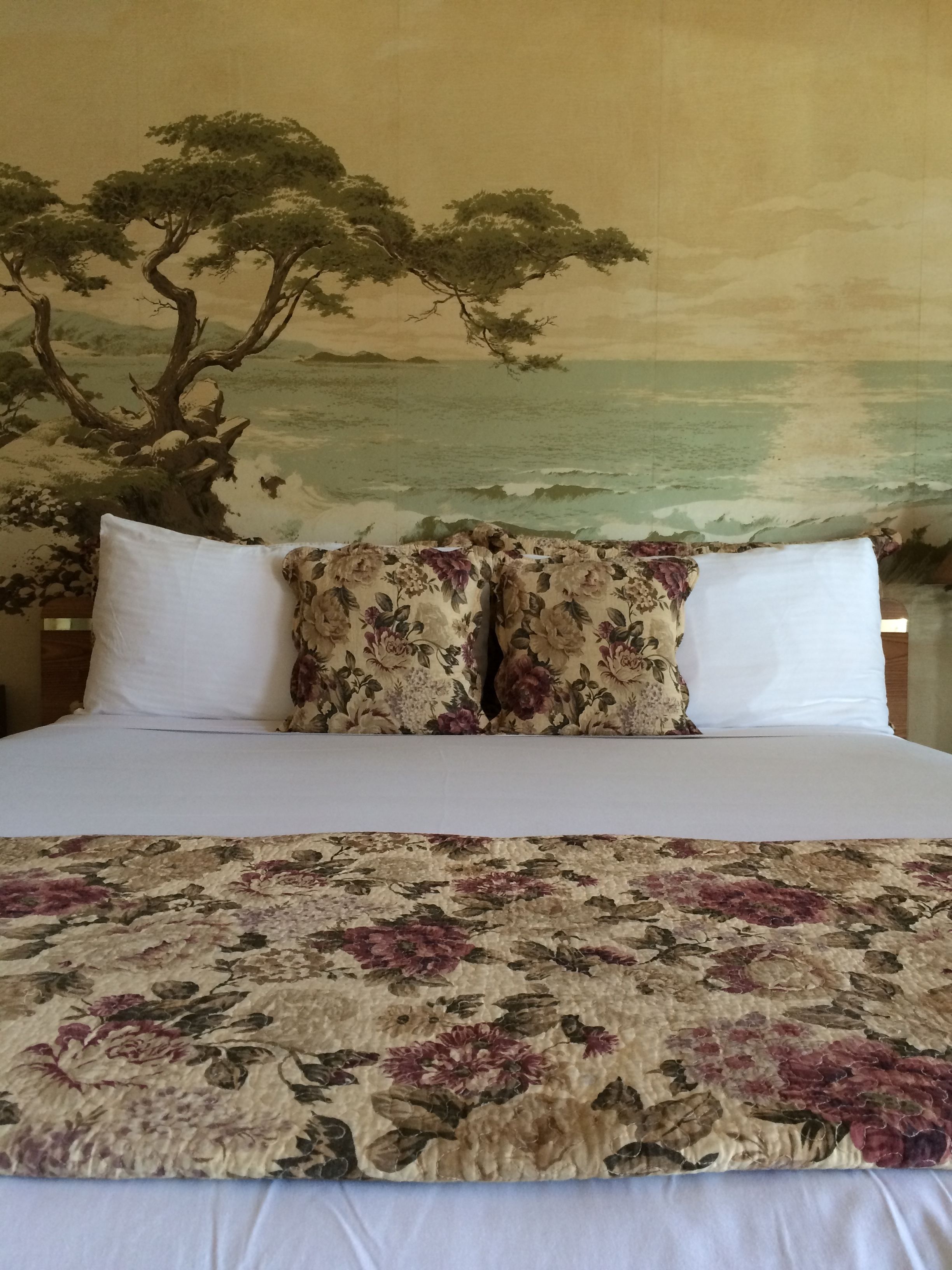 A bed with floral bedding, two white pillows, and a painting of a tree and landscape on the wall behind.