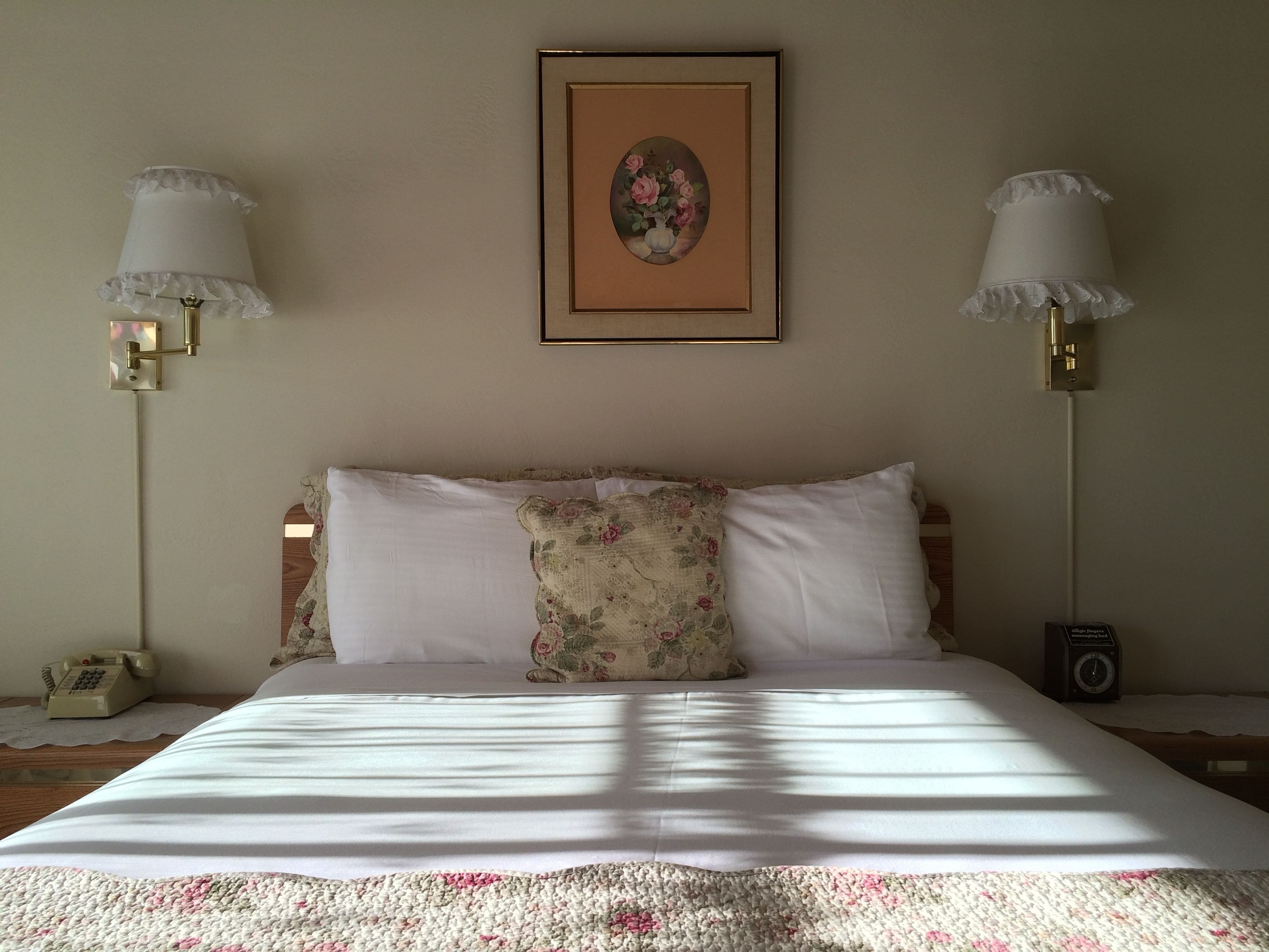 A neatly made bed with a floral pillow, two bedside lamps, a framed picture on the wall, and a vintage radio on the nightstand.