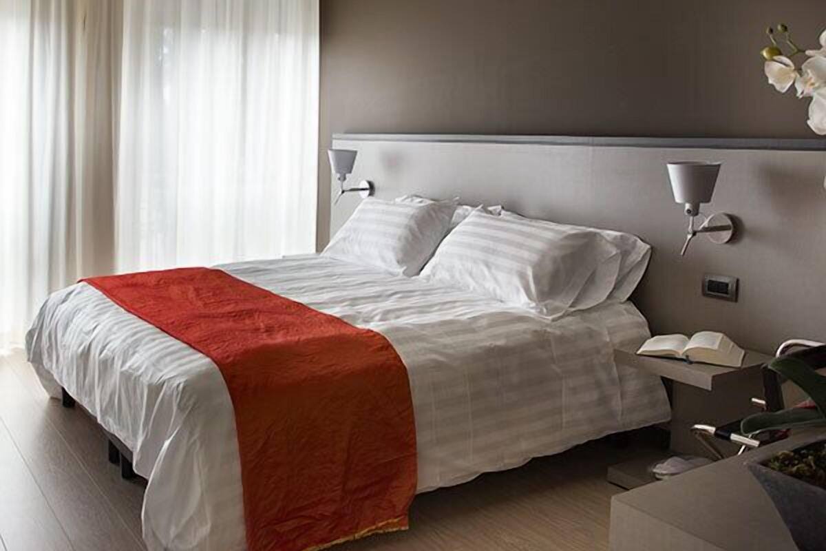A modern hotel room with a large bed, a bedside table with a lamp, and a view of the outdoors through sheer curtains.