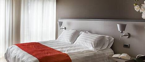 Una habitación de hotel moderna con una cama grande, una mesita de noche con lámpara y vistas al exterior a través de cortinas translúcidas.