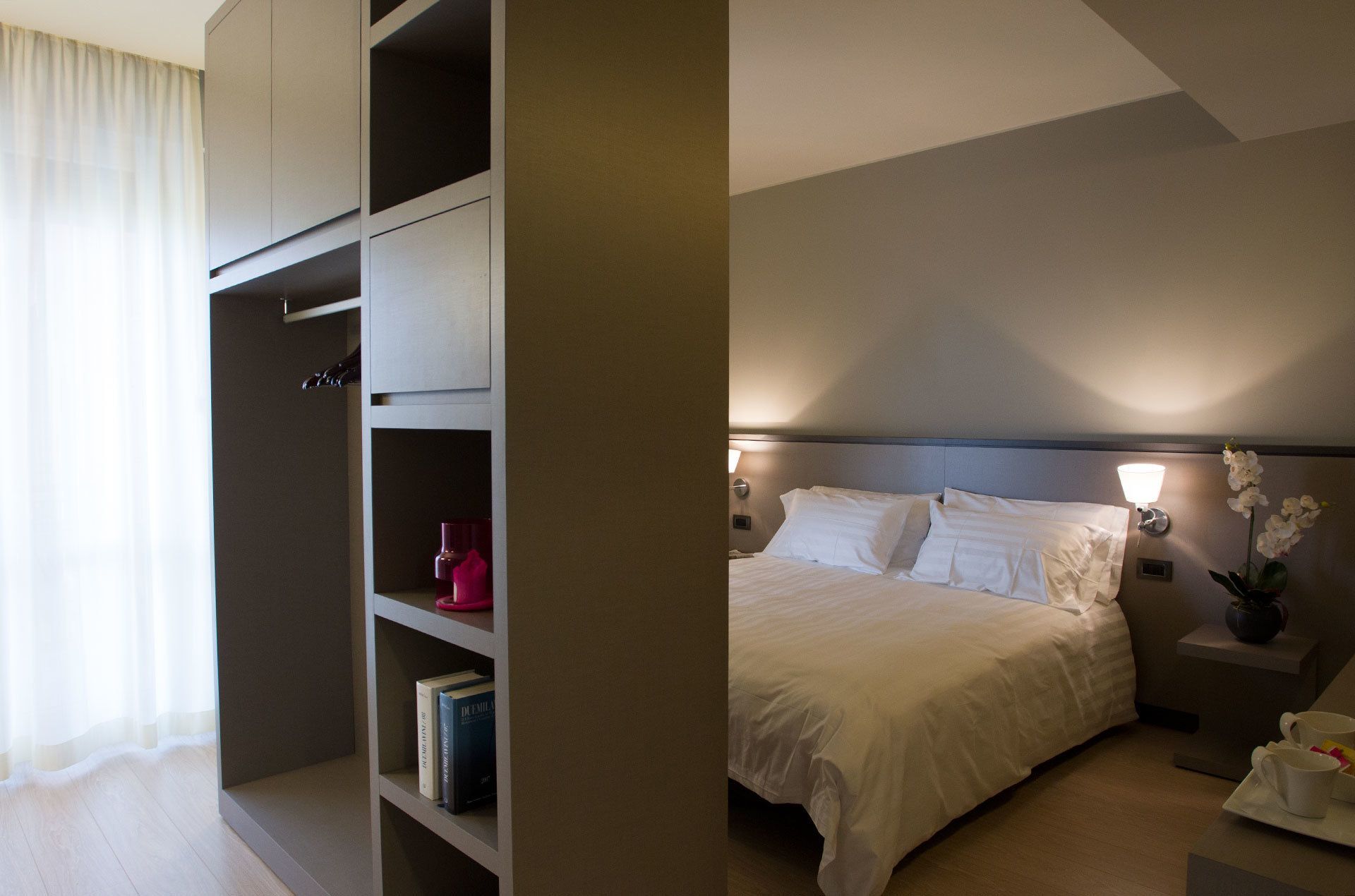 Ein Hotelzimmer mit Bett, Nachttisch, Bücherregal und einem Fenster mit Vorhängen.