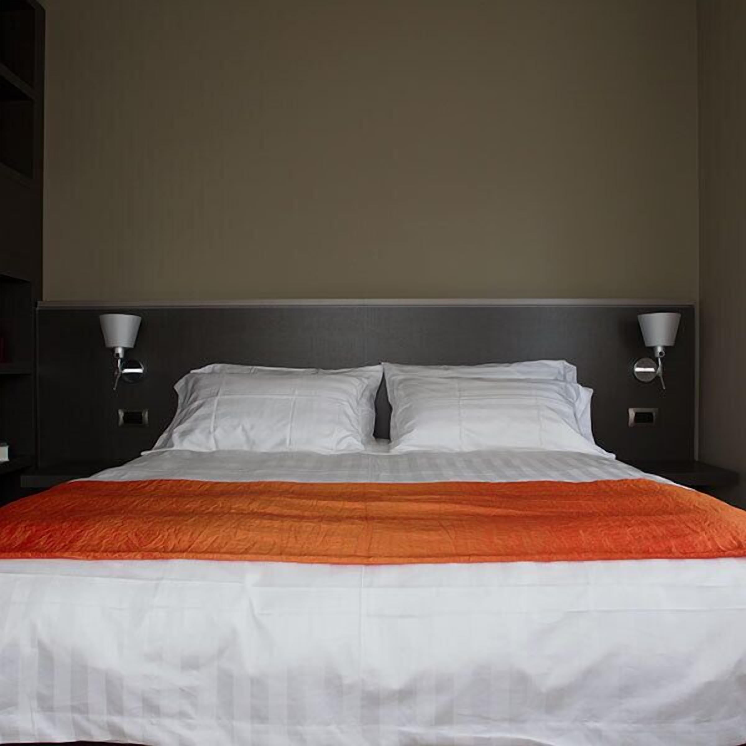 Un lit bien fait, avec une literie blanche et orange, flanqué de deux lampes fixées au mur.