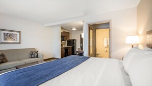 Quarto de hotel com cama, sofá, escrivaninha e cozinha compacta.