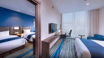 Ein Hotelzimmer mit zwei Betten, einem Schreibtisch mit Stuhl, einem Fernseher und einem großen Fenster mit Vorhängen.