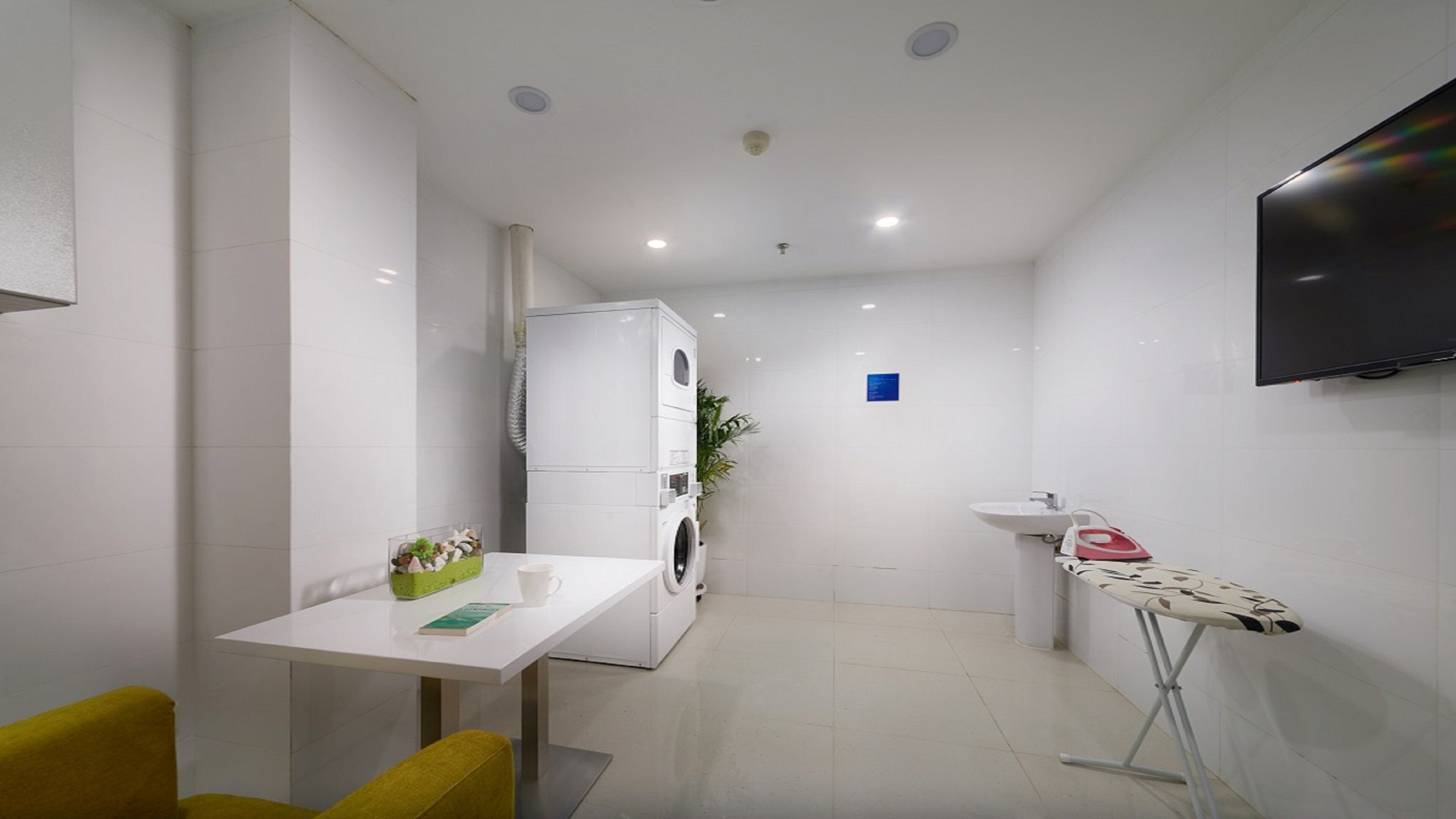 A compact hotel room with a washing machine, a sink, a television, and a small table with a chair.