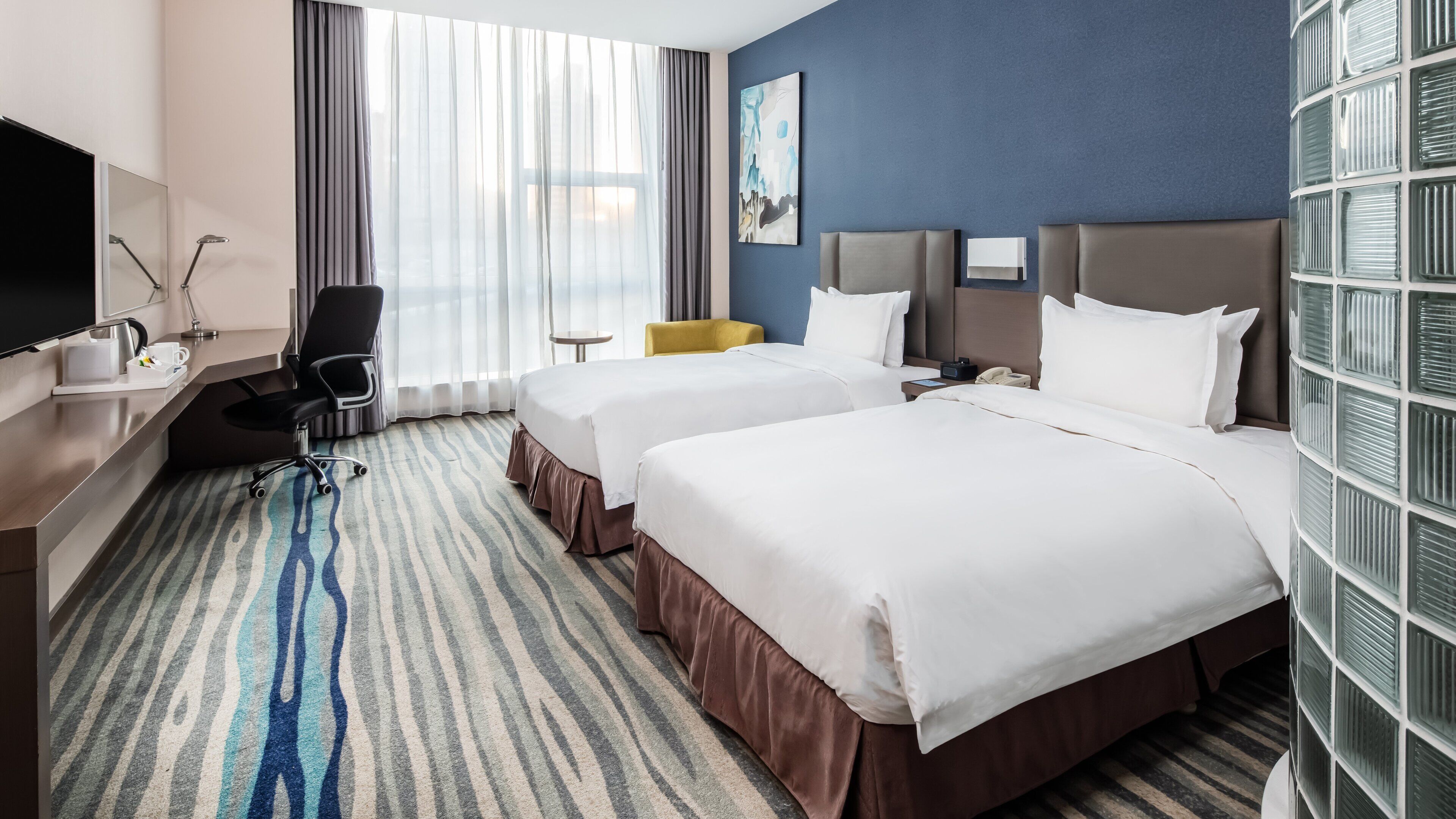 A hotel room with two beds, a desk with a chair, a television, and a large window with curtains.