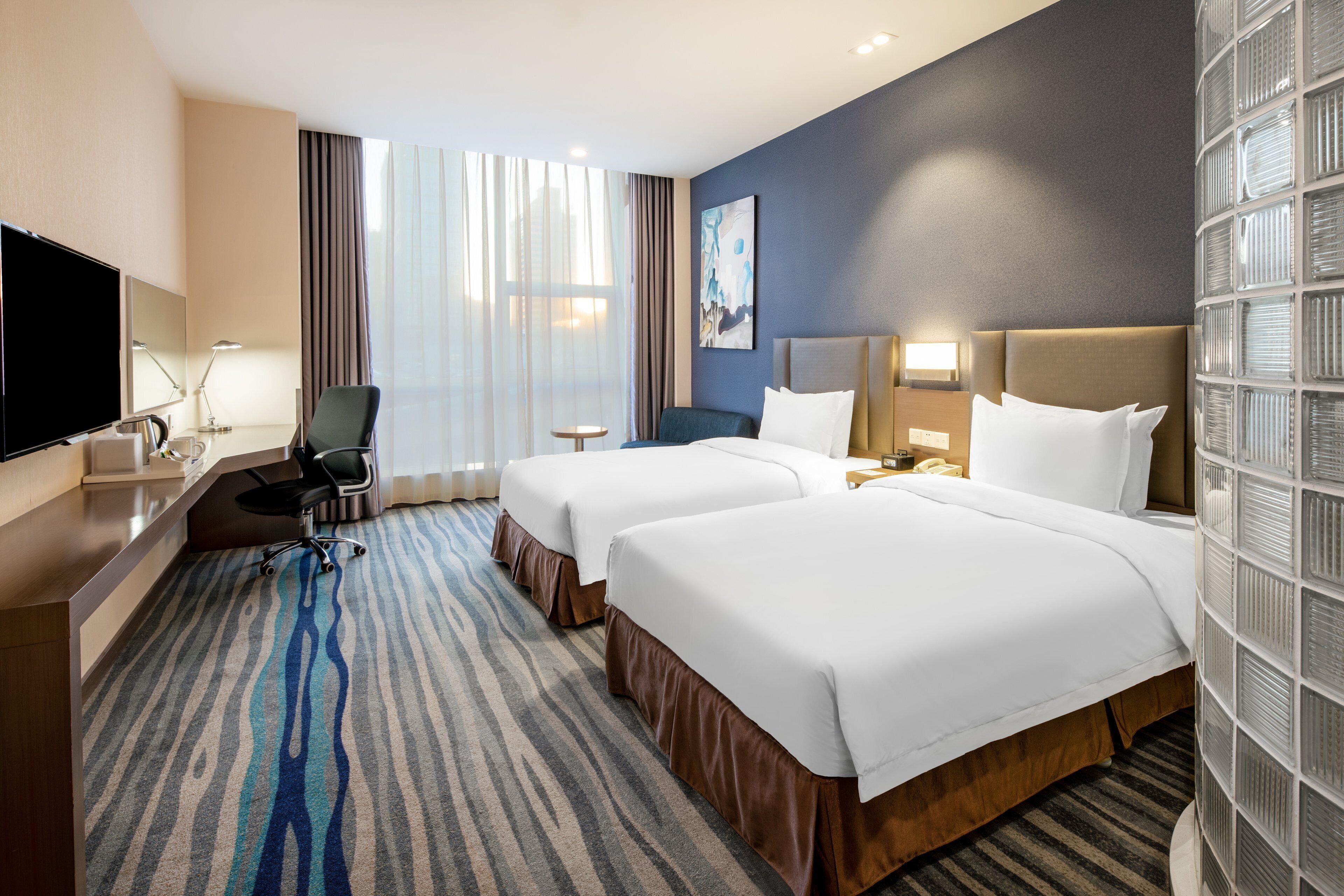 A hotel room with two beds, a desk, a chair, a TV, and a large window with curtains.