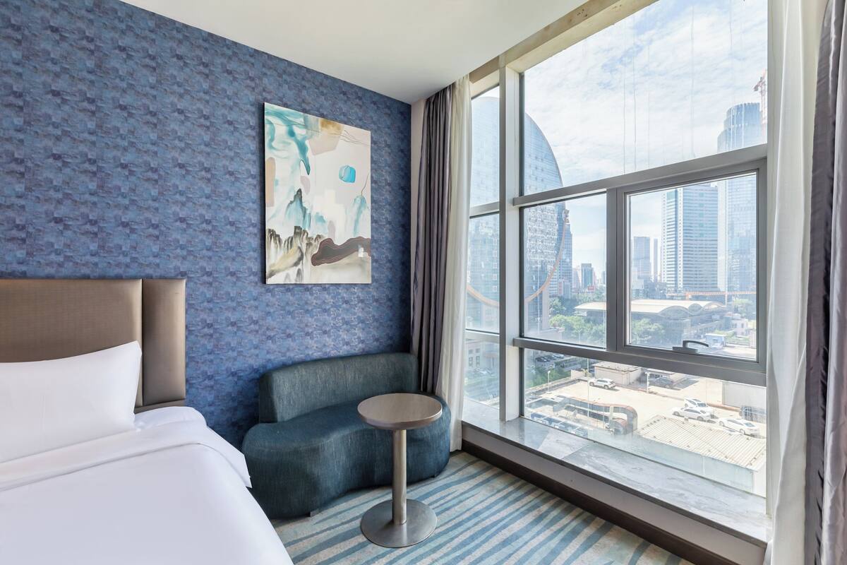 A hotel room with a large window, a bed, a blue sofa, and a small table.