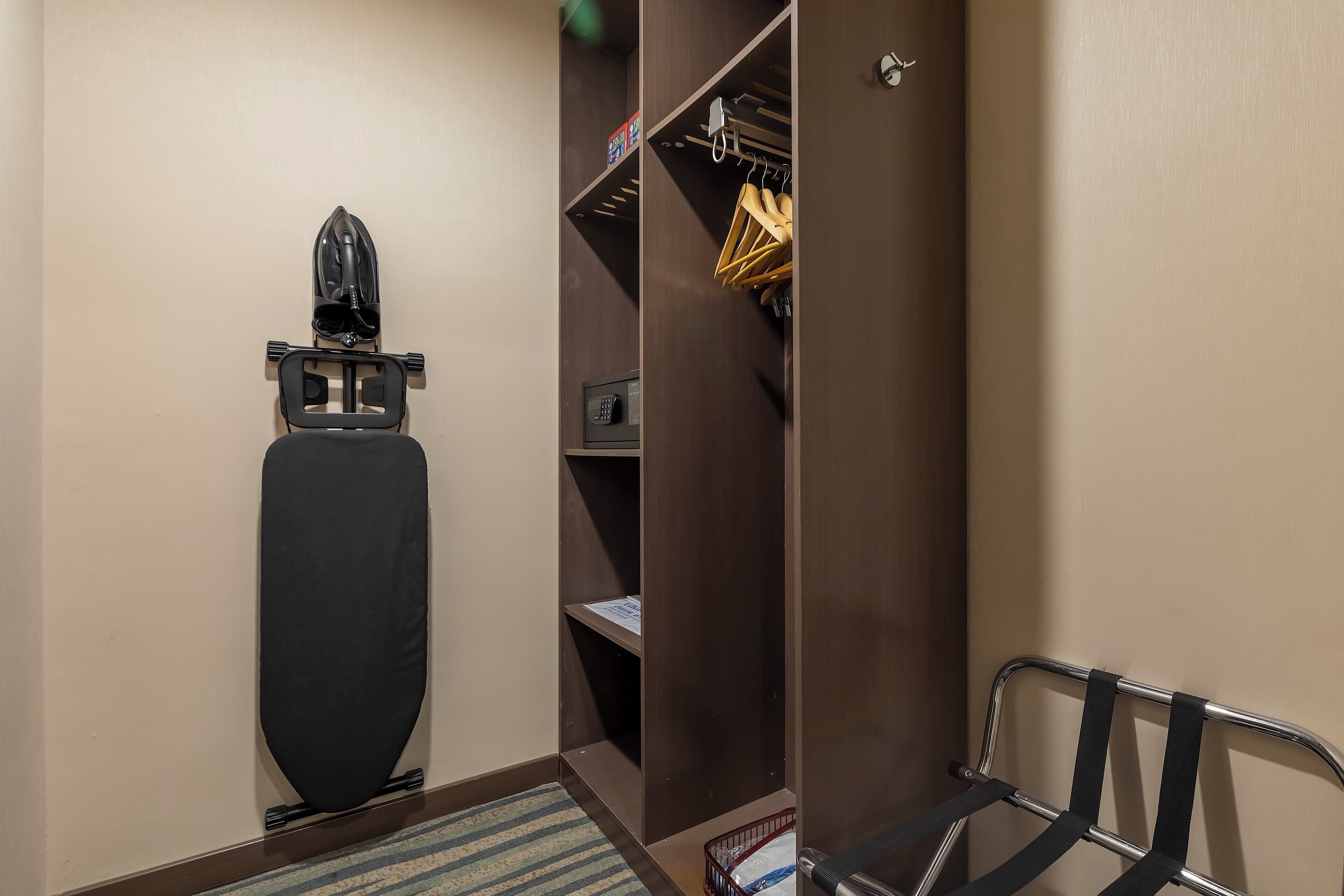 A hotel room with a compact layout, including a bed, a small desk, a closet with hanging clothes, and a shoe rack.