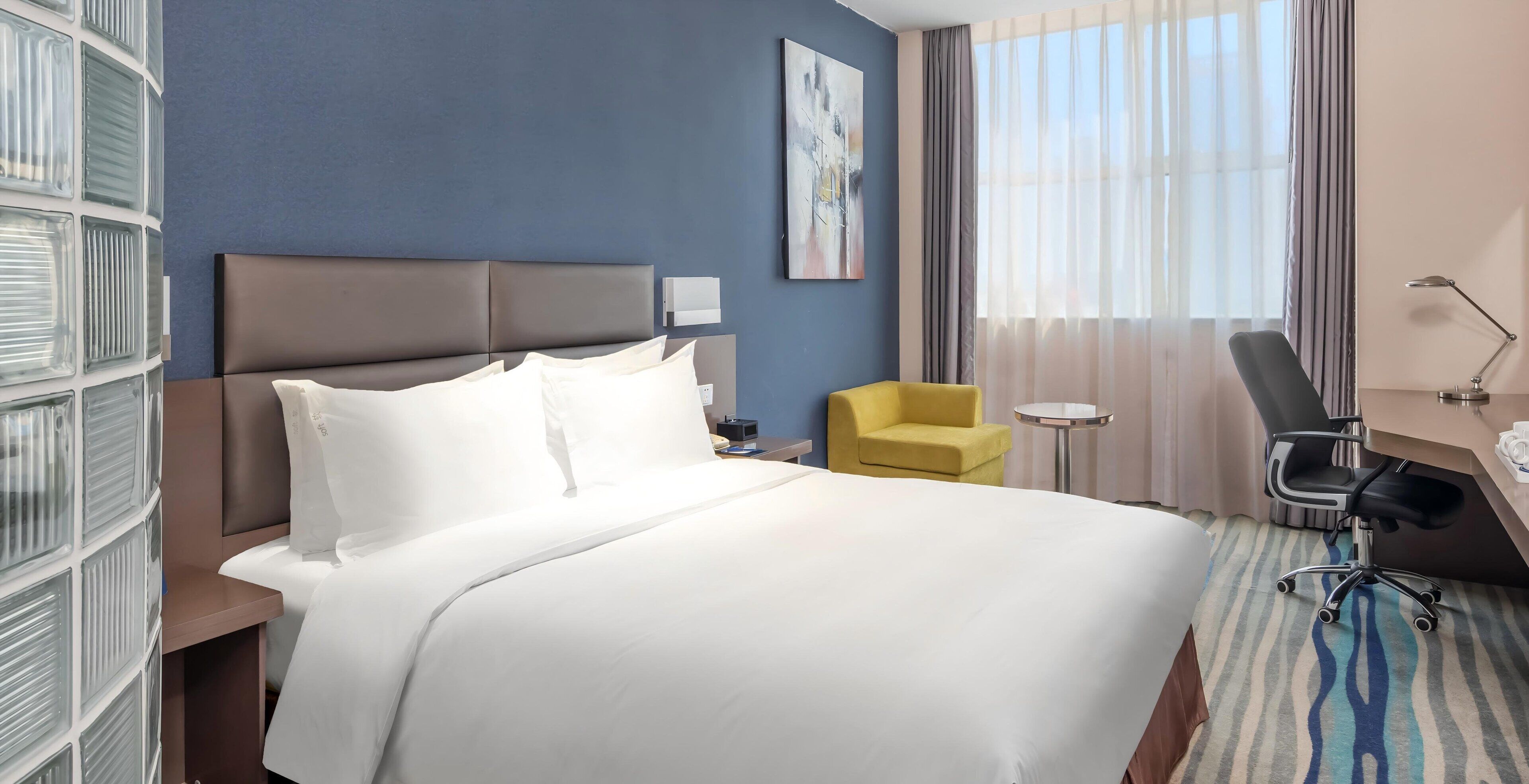 A hotel room with a large bed, a yellow armchair, a desk with a chair, a small round table, and a large window with curtains.