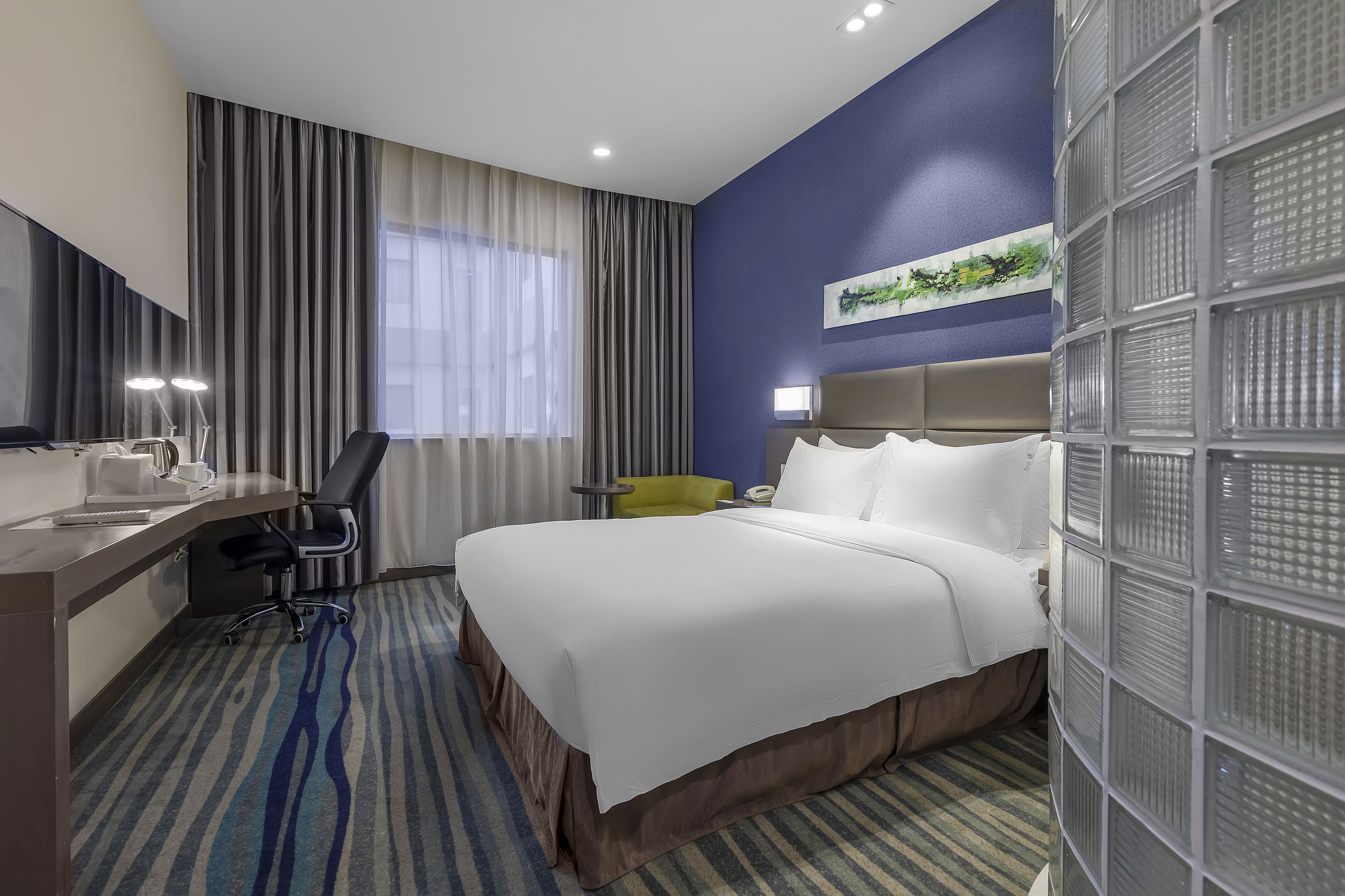 A hotel room with a bed, a desk with a chair, a large window with curtains, and a wall with a decorative panel.