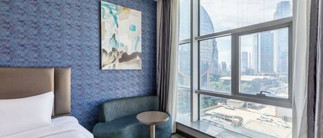 Una habitación de hotel con un ventanal grande, una cama, un sofá azul y una mesita.