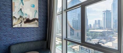 A modern hotel room with a large window offering a city view, a comfortable seating area, and a large abstract painting on the wall.