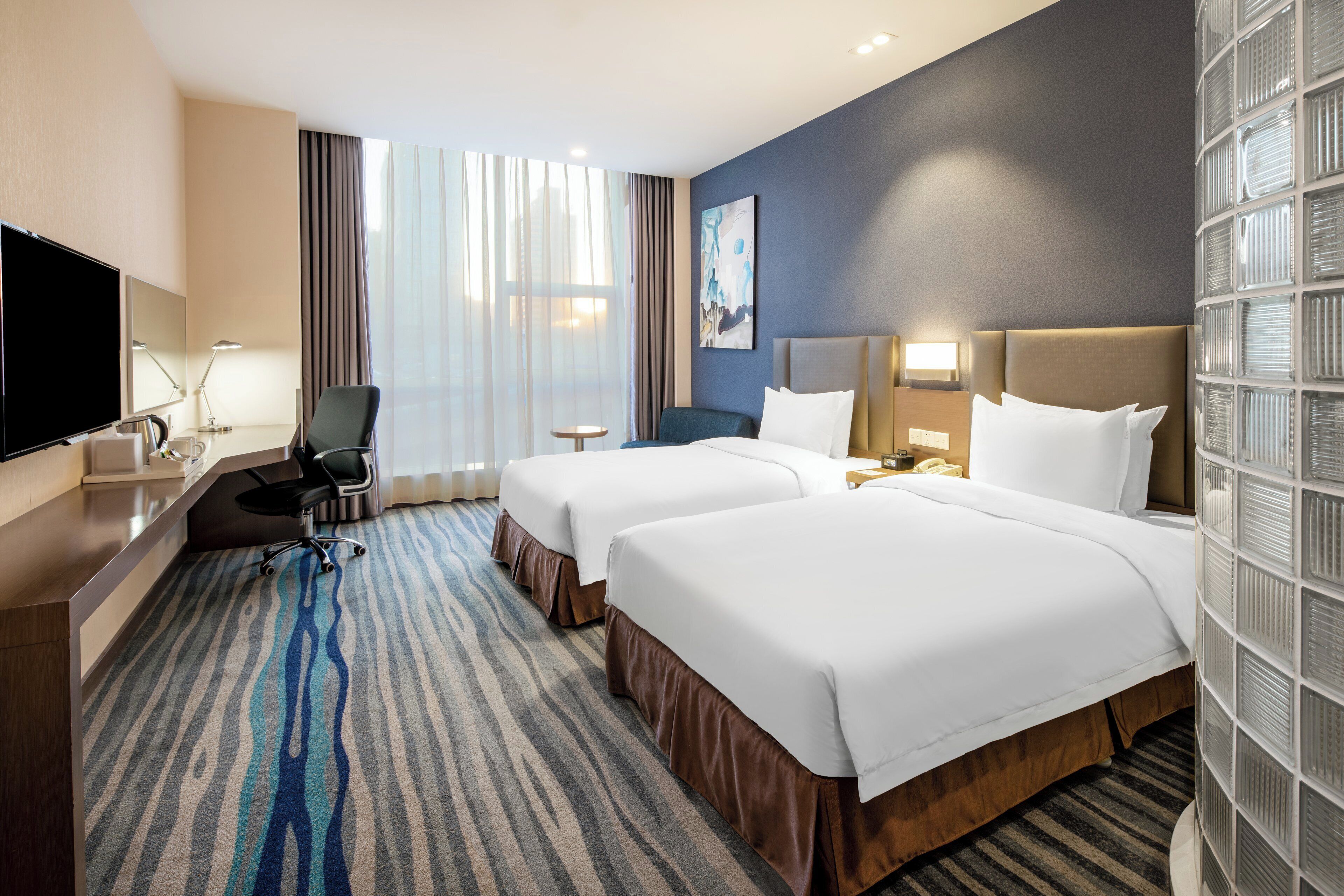 A hotel room with two beds, a desk, a chair, a TV, and a large window with curtains.