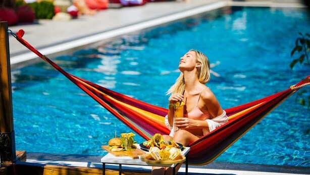 Una persona descansando en una hamaca junto a una piscina, con una mesa de comida y bebidas.