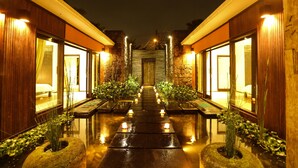 Un pasillo bien iluminado con suelo reflectante que conduce a una puerta central, bordeado por paredes de madera y plantas en macetas.