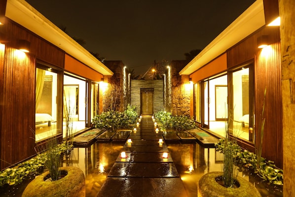 A well-lit corridor with a reflective floor leading to a central door, flanked by wooden walls and potted plants.