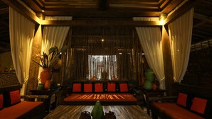 Una habitación con techo de madera, un ventanal con cortinas, un sofá con cojines rojos y una mesa con un jarrón y dos macetas.