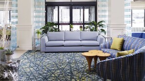 A spacious lobby with a large blue patterned rug, a grey sofa, and a blue armchair. There are hanging white pendant lights and a large window with a view. - Hilton Garden Inn Longview (Longview)