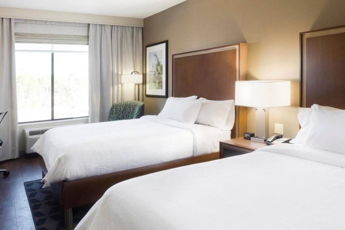 Une chambre d’hôtel avec deux lits, une chaise, un bureau et une fenêtre avec des rideaux.