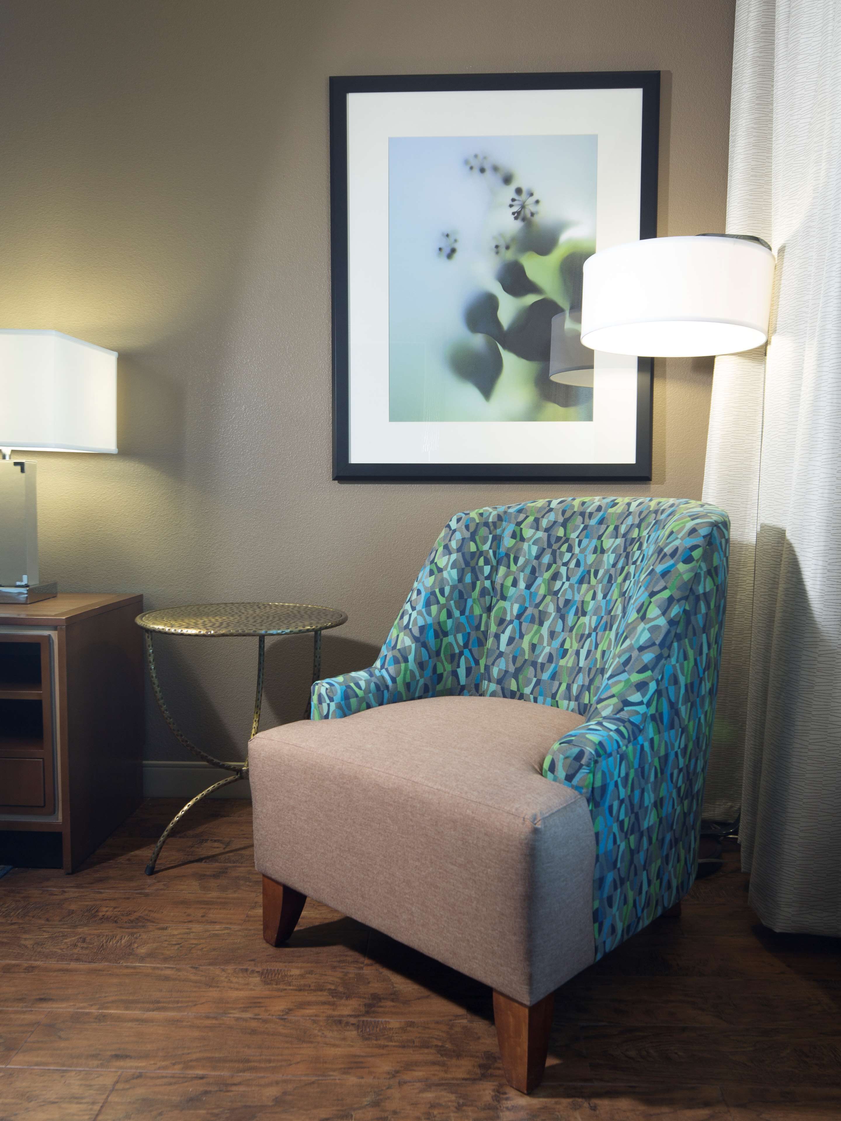 A room with a patterned armchair, a framed abstract painting, a small round table, and a lamp.