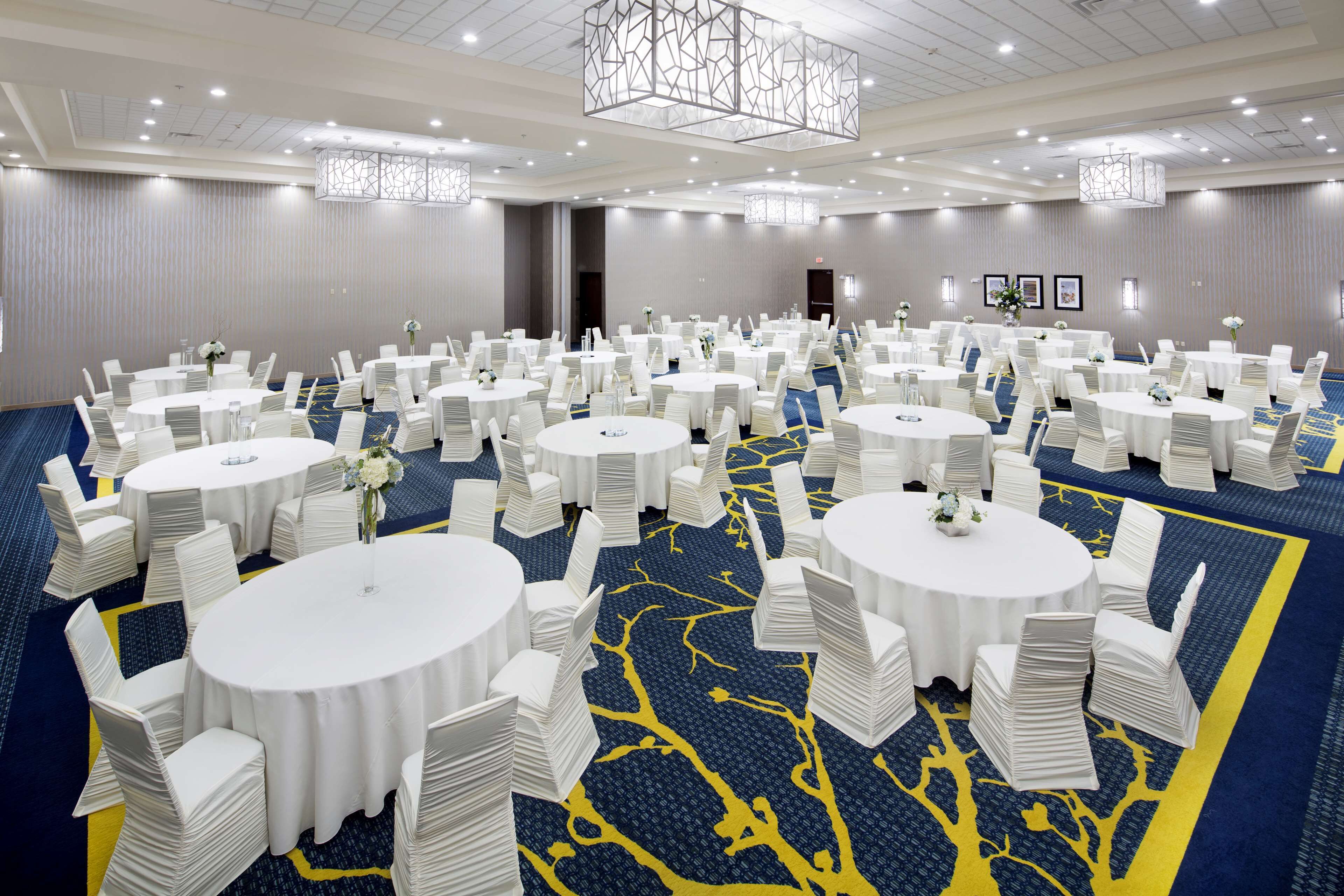 Uma grande sala de conferências com várias mesas redondas, cada uma rodeada por cadeiras brancas e pequenos arranjos florais. A sala possui um tapete azul com um design de árvore amarela, e o teto tem um candelabro geométrico.