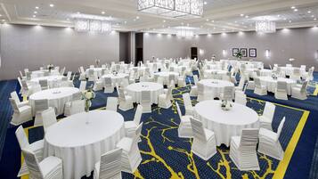 Uma grande sala de conferências com várias mesas redondas, cada uma rodeada por cadeiras brancas e pequenos arranjos florais. A sala possui um tapete azul com um design de árvore amarela, e o teto tem um candelabro geométrico.