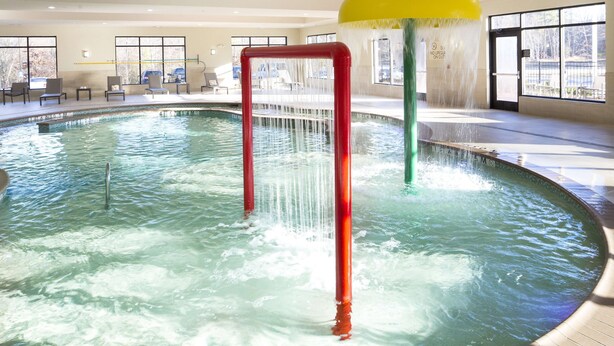 An indoor pool with clear water, a red and yellow water feature, and a seating area with chairs and tables. - Hilton Garden Inn Longview (Longview)