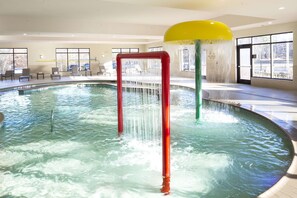 An indoor pool with clear water, a red and yellow water feature, and a seating area with chairs and tables.