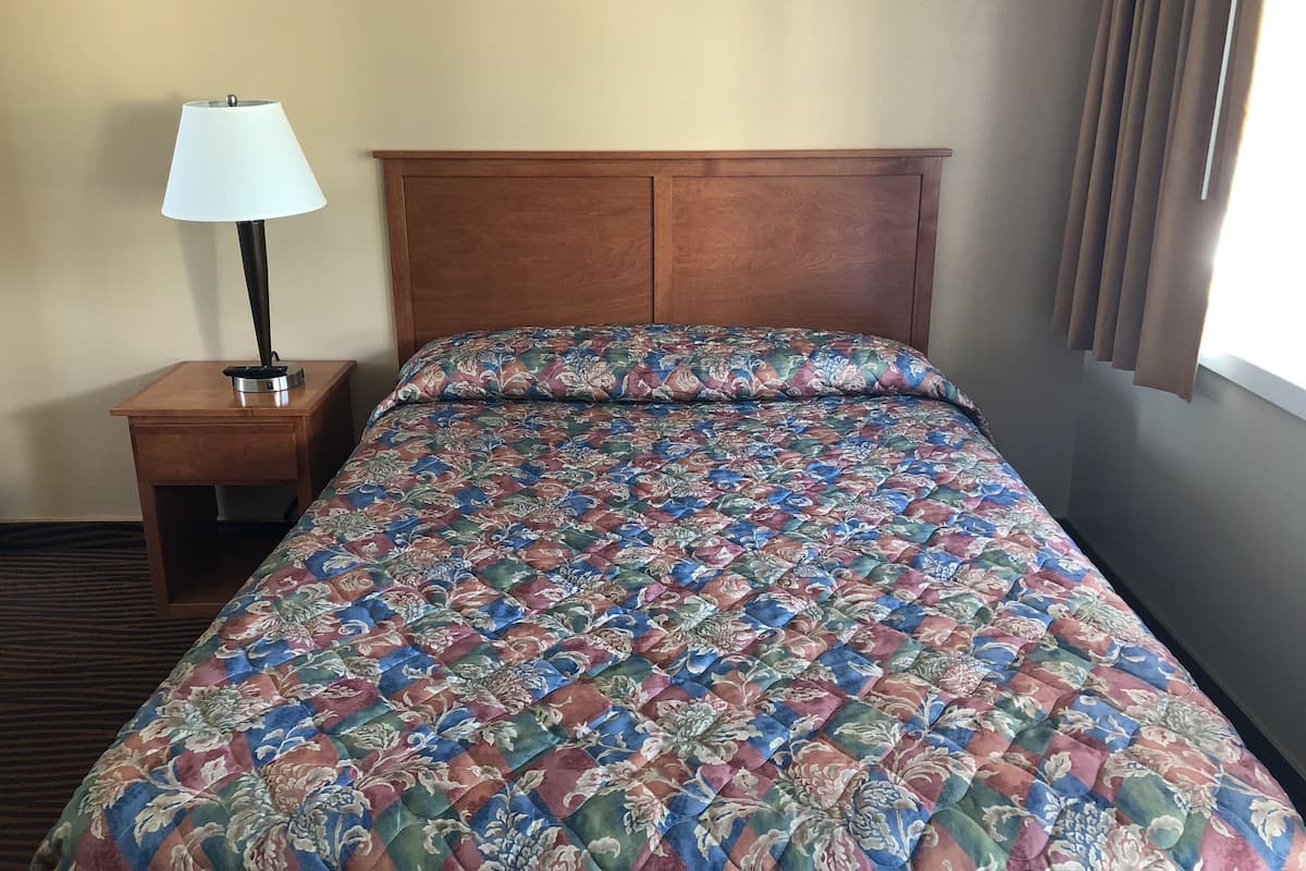 A hotel room with a wooden headboard, a floral-patterned bedspread, a nightstand with a lamp, and a window with curtains.