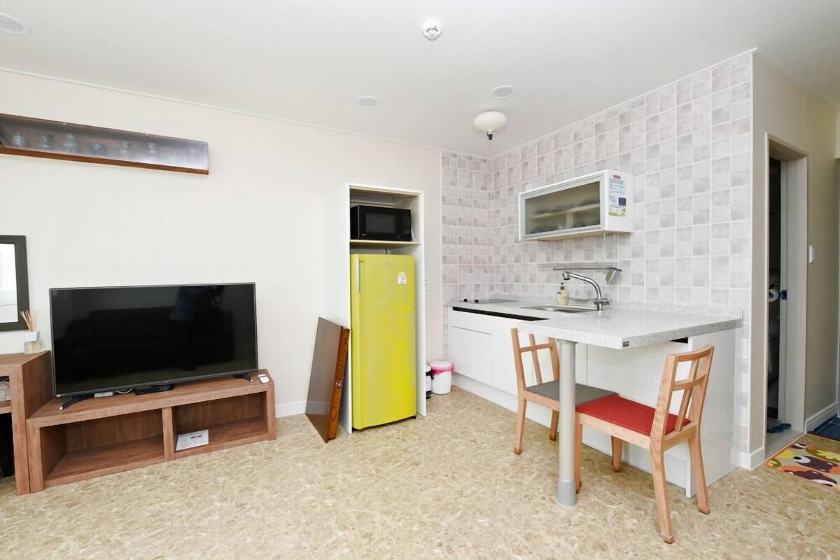 A compact living space with a kitchenette, a flat-screen TV on a wooden stand, a yellow refrigerator, and a dining area with a chair.
