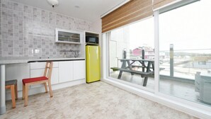 Una cocina moderna con armarios blancos, un microondas y un refrigerador amarillo. Hay una silla de madera y un banco rojo. Un ventanal ofrece vistas a un balcón con una mesa de picnic.